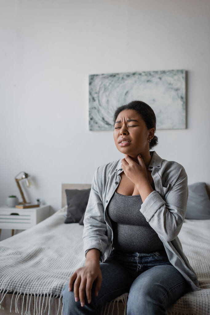 Estressado multirracial mulher tocando dor de garganta enquanto sentado na cama em casa  - Foto, Imagem