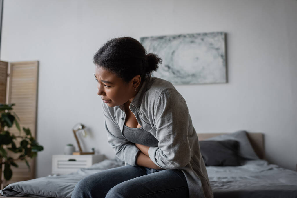 Moe multiraciale vrouw gevoel kramp in de maag tijdens het zitten op bed  - Foto, afbeelding
