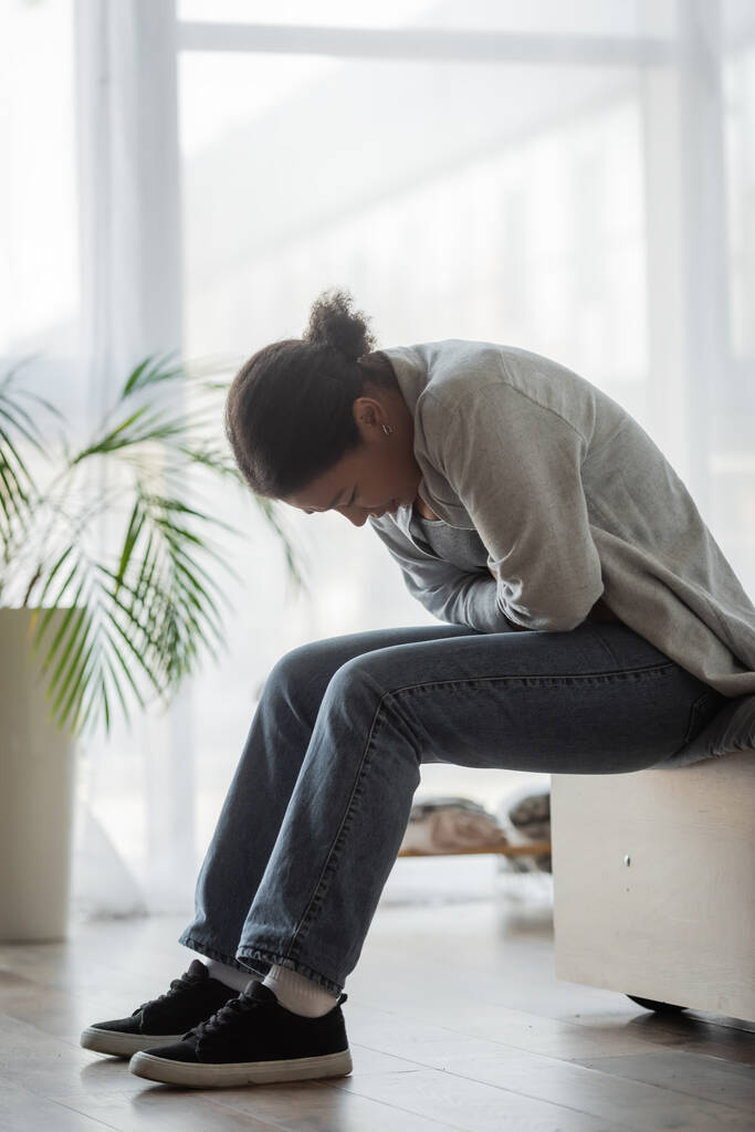 Λυπημένη πολυφυλετική γυναίκα αισθάνεται άσχημα, ενώ κάθεται στο κρεβάτι στο σπίτι - Φωτογραφία, εικόνα