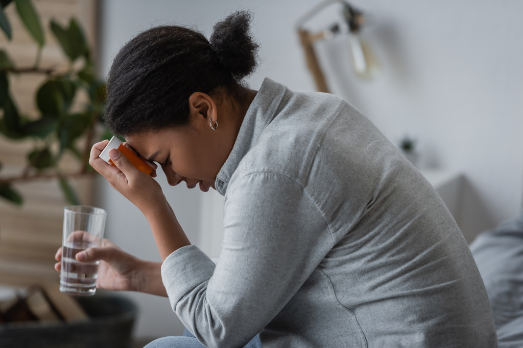Boční pohled na nespokojenost multiracial žena drží pilulky a vodu v ložnici  - Fotografie, Obrázek