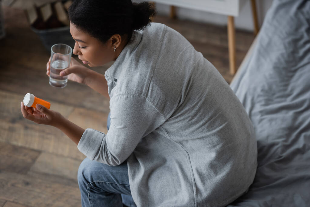 Yleisnäkymä surullinen monirotuinen nainen katselee pillereitä ja tilalla lasi vettä makuuhuoneessa  - Valokuva, kuva