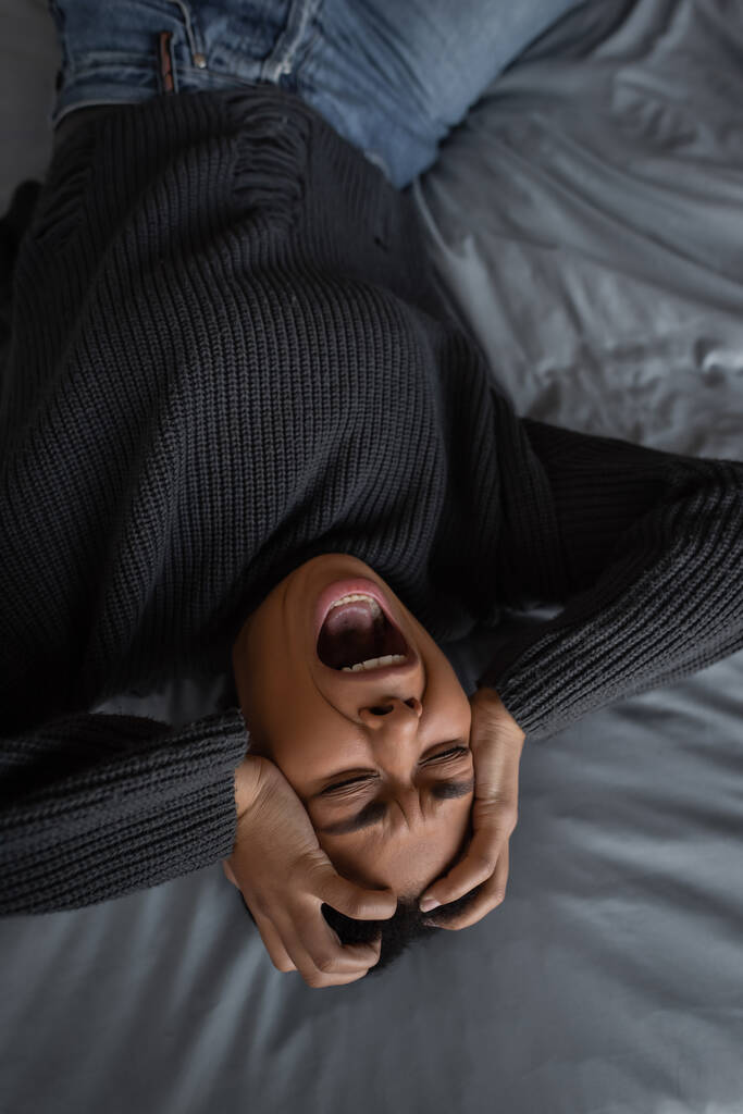 Visão superior da mulher multirracial com depressão gritando enquanto deitado na cama  - Foto, Imagem
