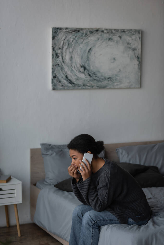 mujer multirracial con ansiedad hablando en el teléfono inteligente en la cama en el dormitorio  - Foto, Imagen