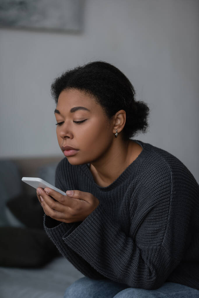 Mladý multiracial žena pomocí smartphone zatímco sedí na rozmazané posteli doma  - Fotografie, Obrázek