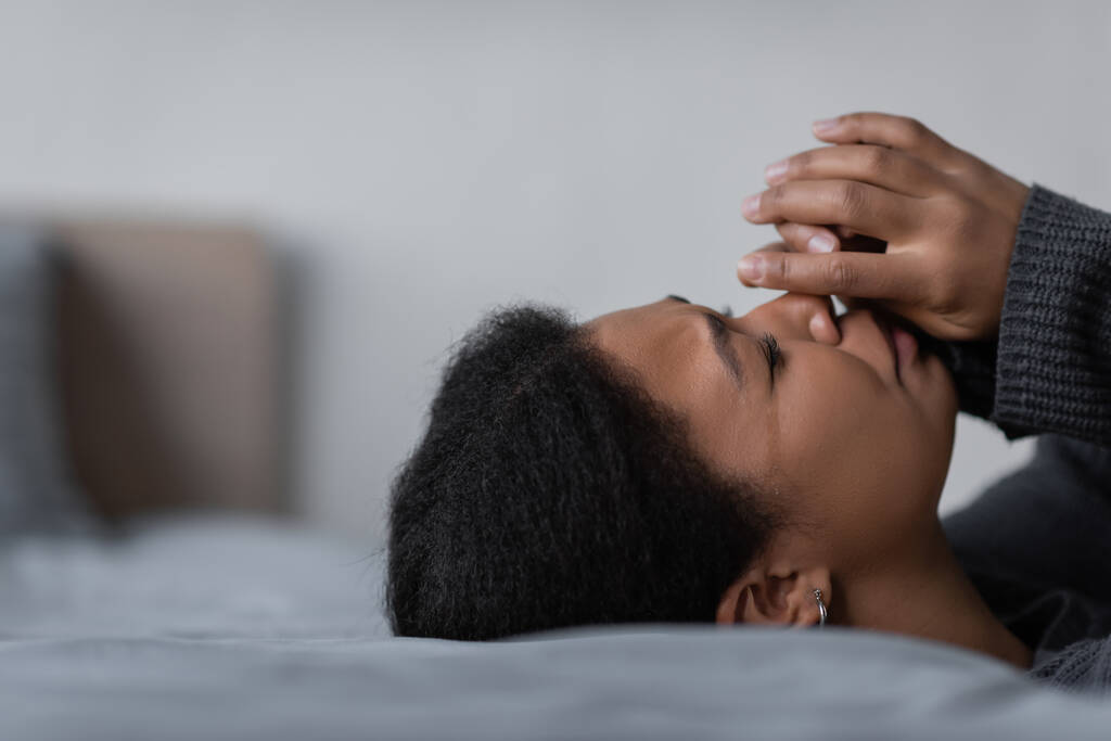 Νεαρή πολυφυλετική γυναίκα με κατάθλιψη κλαίει ενώ βρίσκεται στο κρεβάτι στην κρεβατοκάμαρα  - Φωτογραφία, εικόνα