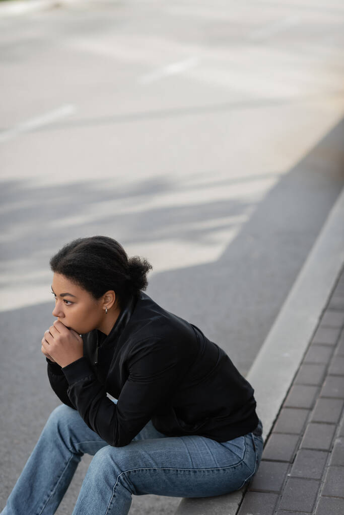 Mujer joven multirracial con depresión mirando hacia otro lado mientras está sentada en la frontera al aire libre  - Foto, Imagen