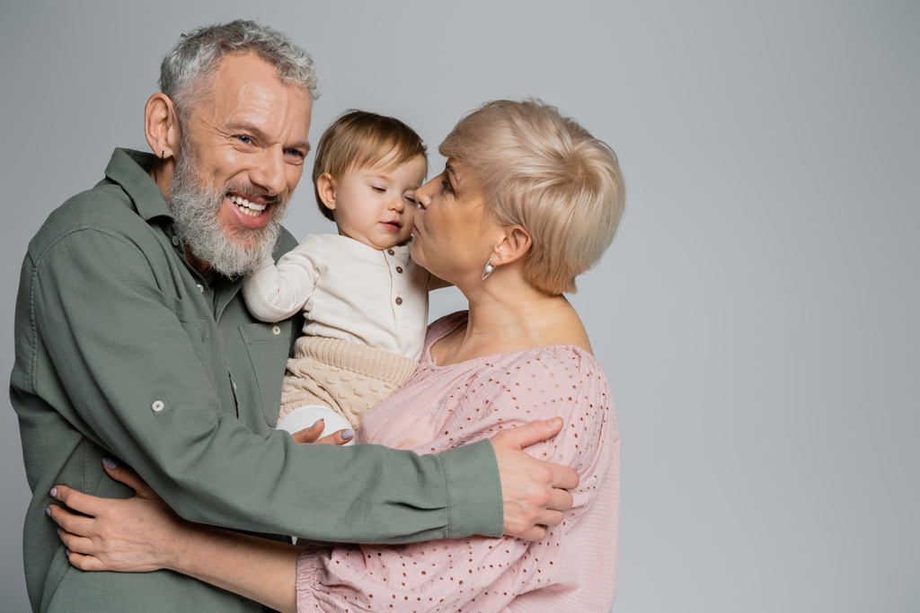 mature woman kissing granddaughter near pleased bearded husband isolated on grey - Photo, Image