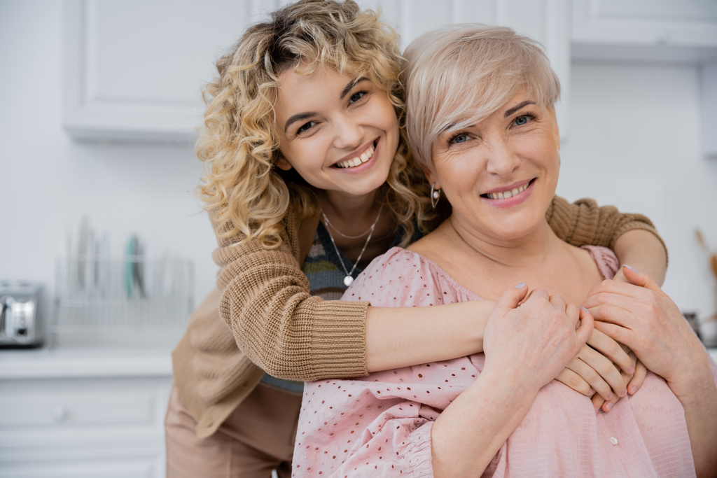 nadšený žena s kudrnaté blond vlasy objímající šťastnou matku a usmívající se na kameru v kuchyni - Fotografie, Obrázek