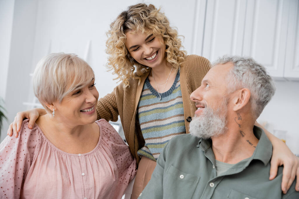 lächelnde Frau mit blonden welligen Haaren umarmt fröhliche Eltern mittleren Alters in der heimischen Küche - Foto, Bild