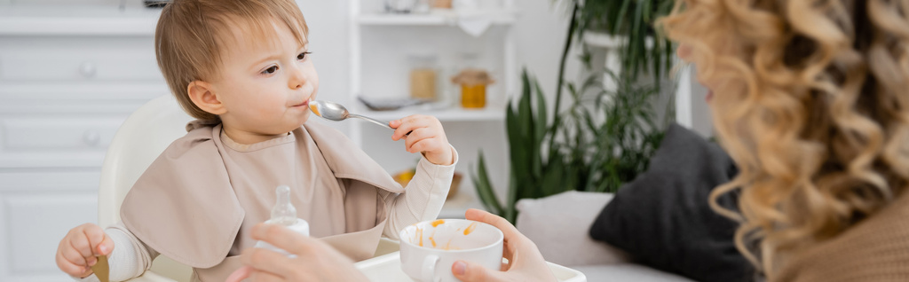 małe dziecko z łyżeczką jedzące puree w pobliżu rozmytej matki trzymające miskę w kuchni, baner - Zdjęcie, obraz