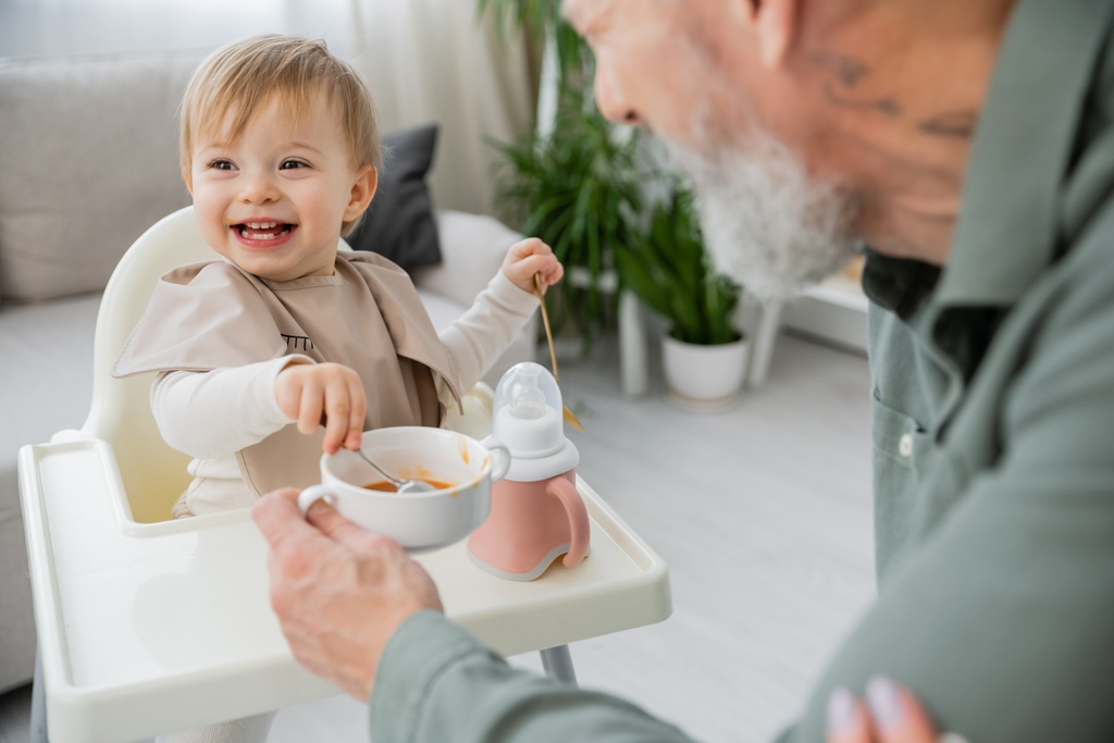 беззаботная девушка с ложками сидя на детском стульчике возле чаши с завтраком и дедушка на размытом переднем плане на кухне - Фото, изображение