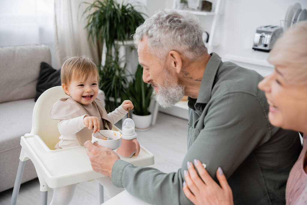 casal maduro sorrindo perto de neta despreocupada com colheres sentadas na cadeira do bebê perto de tigela com café da manhã na cozinha - Foto, Imagem