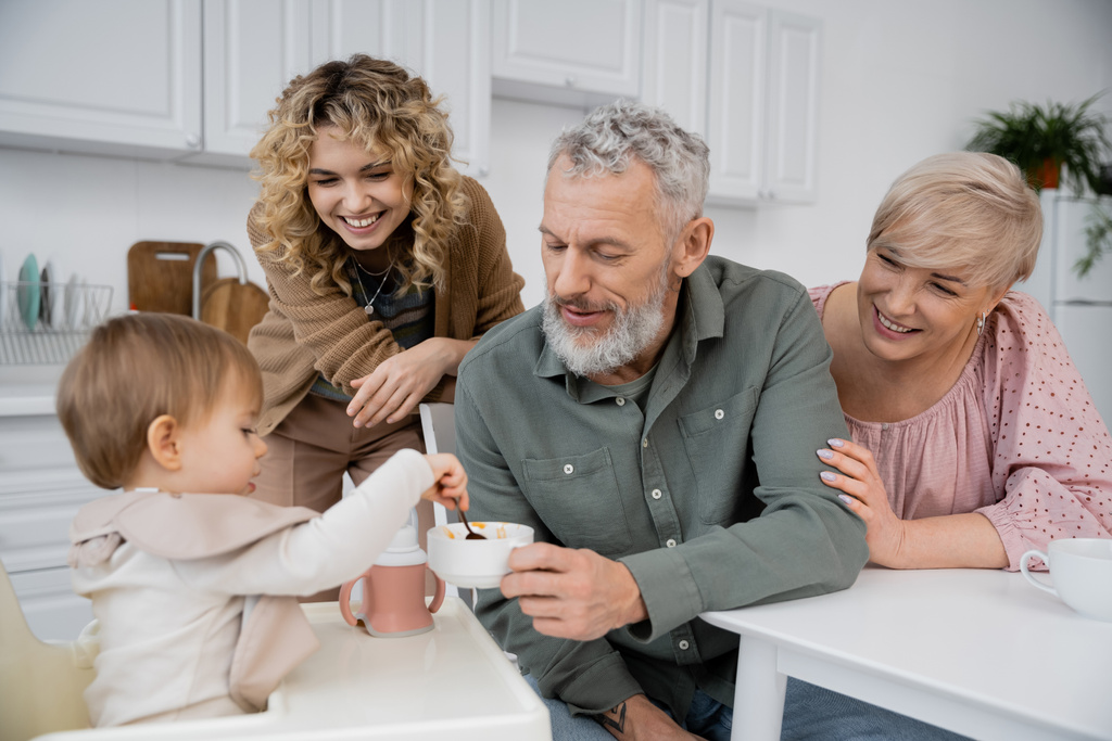 man met baard zitten met kom in de buurt vrolijke familie en peuter kleindochter met lepel in de keuken - Foto, afbeelding