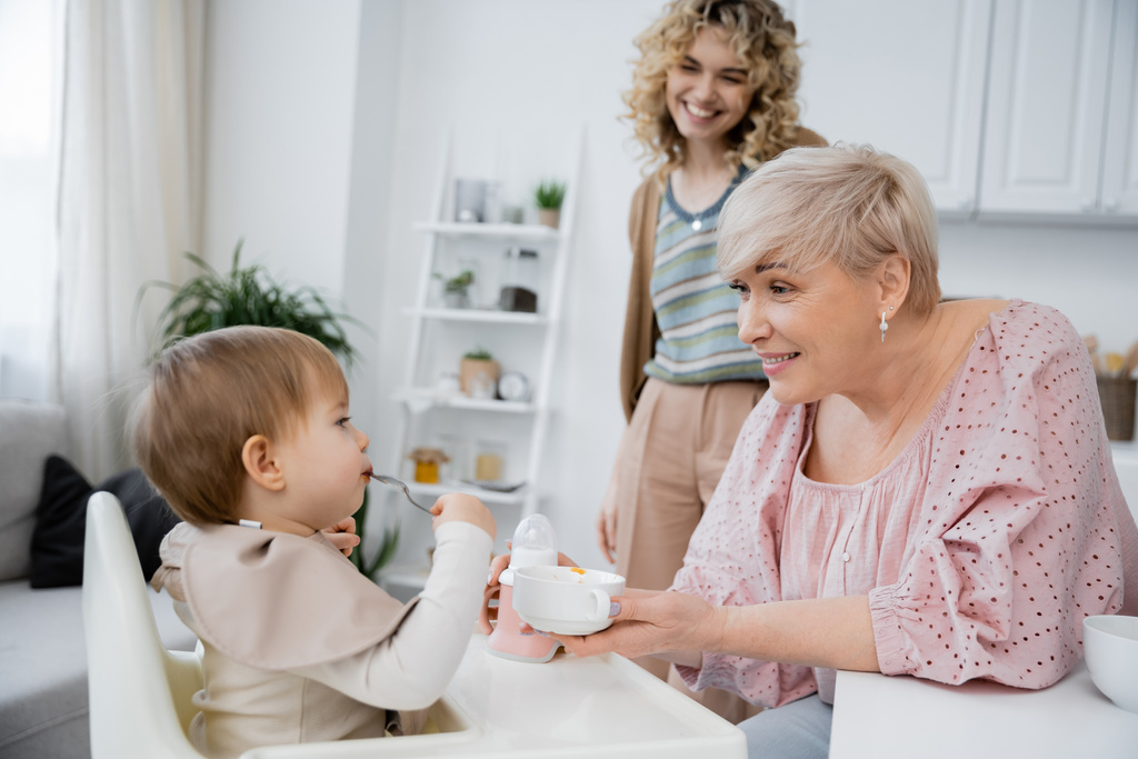 задоволена жінка середнього віку тримає миску біля дитячої онуки з ложкою під час сніданку на кухні
 - Фото, зображення