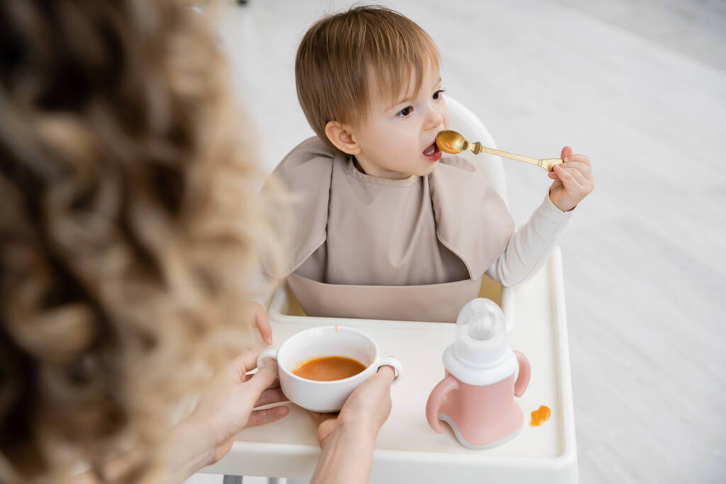 maluch dziewczyna siedzi na dziecko krzesło i jedzenie puree w pobliżu zamazane mama z misa w kuchni - Zdjęcie, obraz