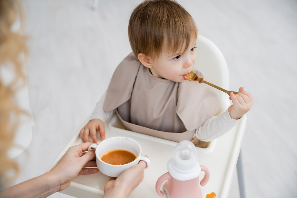 mała dziewczynka z łyżeczką jedząc smaczny puree siedząc w foteliku obok matki z misą w kuchni - Zdjęcie, obraz