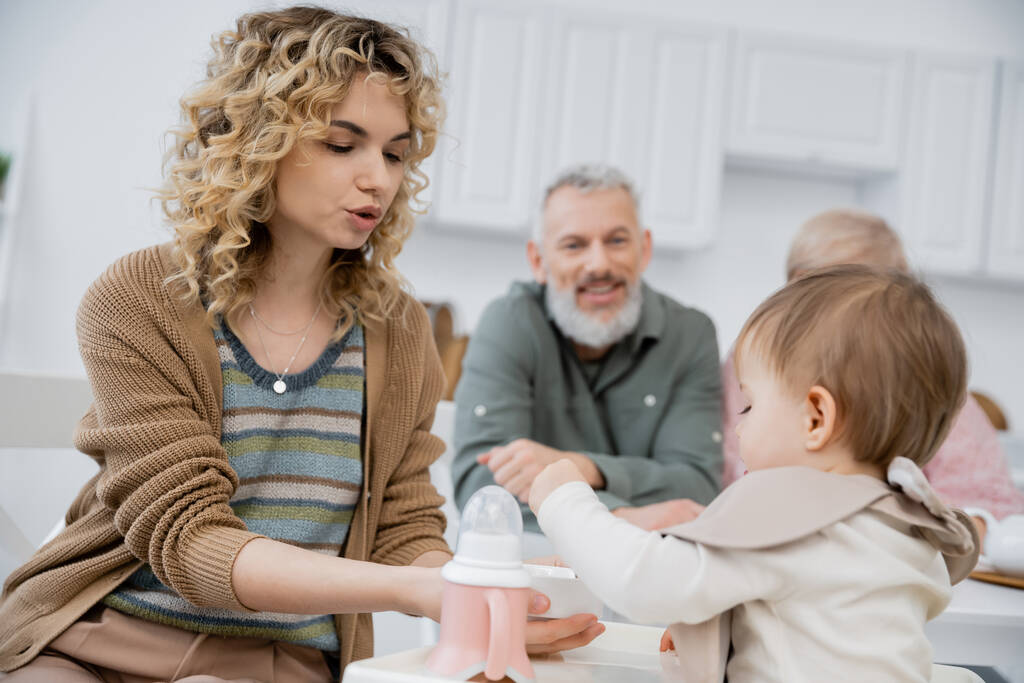 femme nourrissant petite fille avec petit déjeuner près de parents matures souriant sur fond flou dans la cuisine - Photo, image