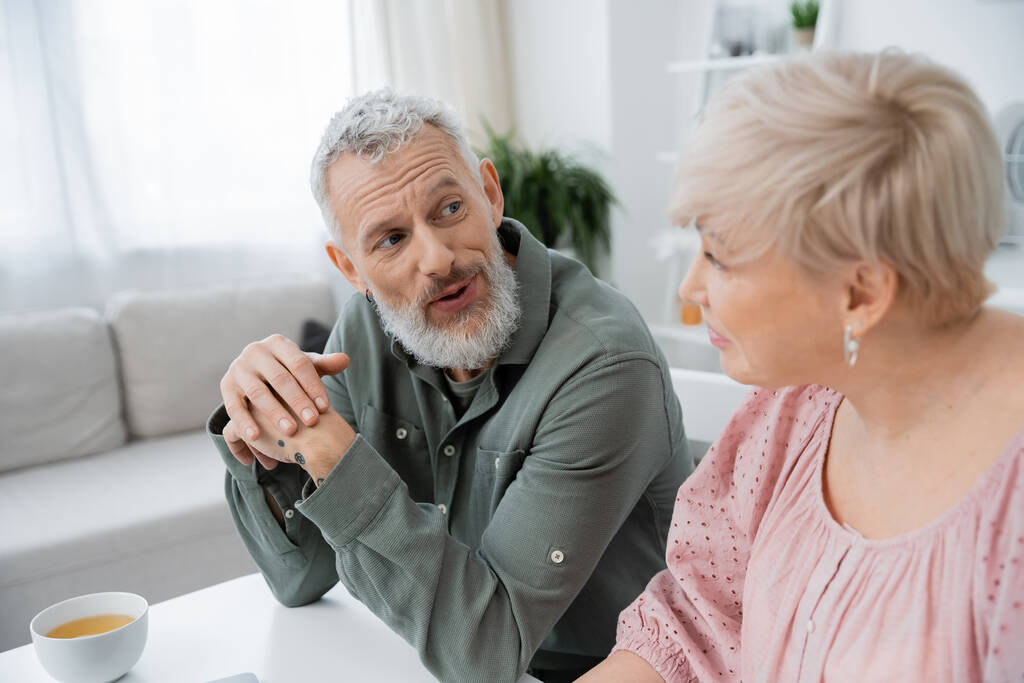 позитивний бородатий чоловік розмовляє з розмитою дружиною середнього віку біля чашки чаю на столі на кухні
  - Фото, зображення