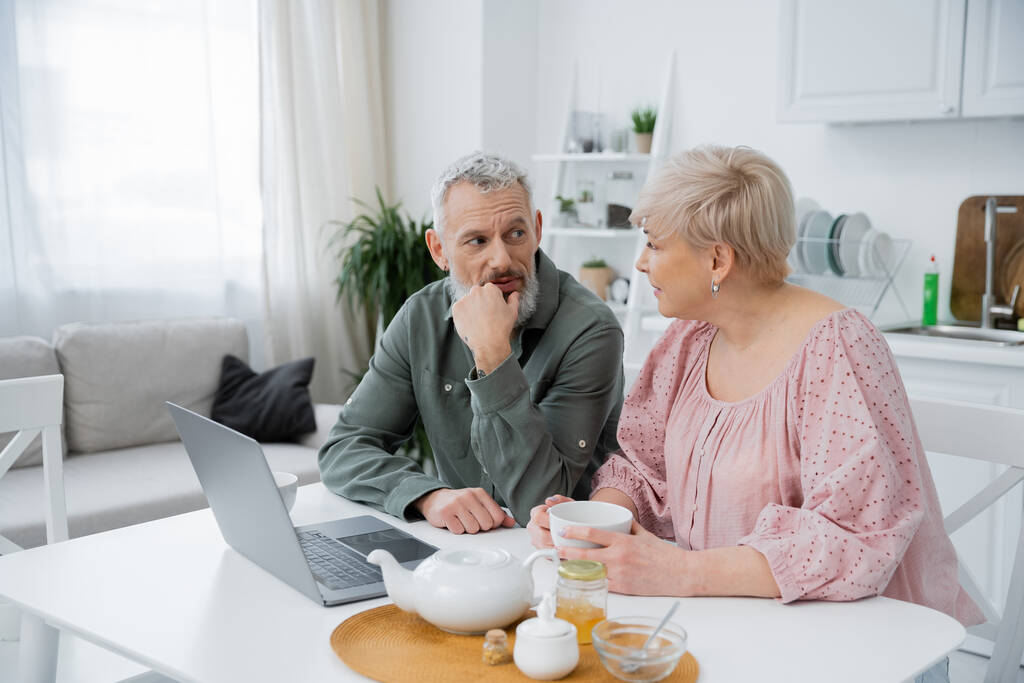barbuto uomo di mezza età guardando moglie mentre parla vicino tazza di tè e laptop sul tavolo in cucina moderna - Foto, immagini