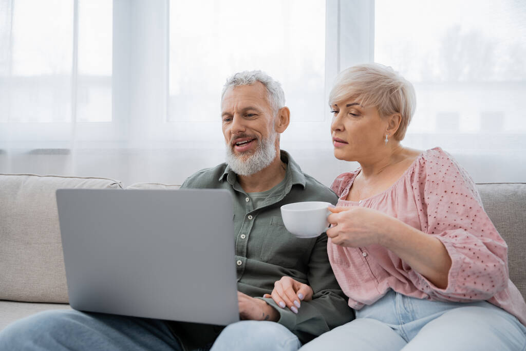 kobieta w średnim wieku trzyma filiżankę herbaty podczas oglądania filmu na laptopie w pobliżu uśmiechniętego męża na kanapie w salonie - Zdjęcie, obraz