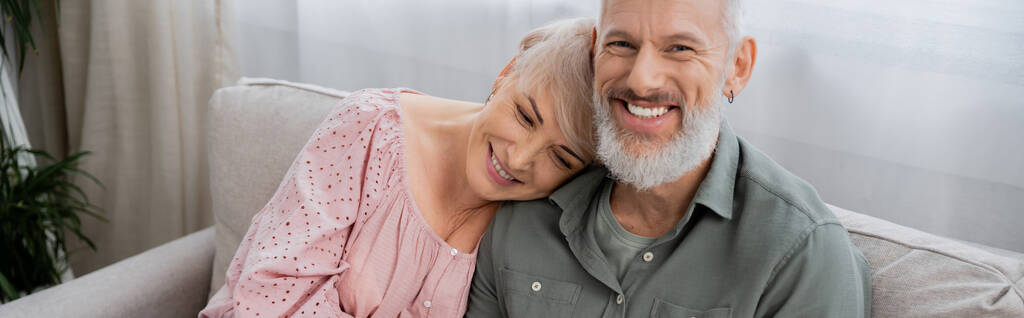 spensierato uomo di mezza età che sorride alla telecamera vicino alla moglie felice sul divano in soggiorno, banner - Foto, immagini