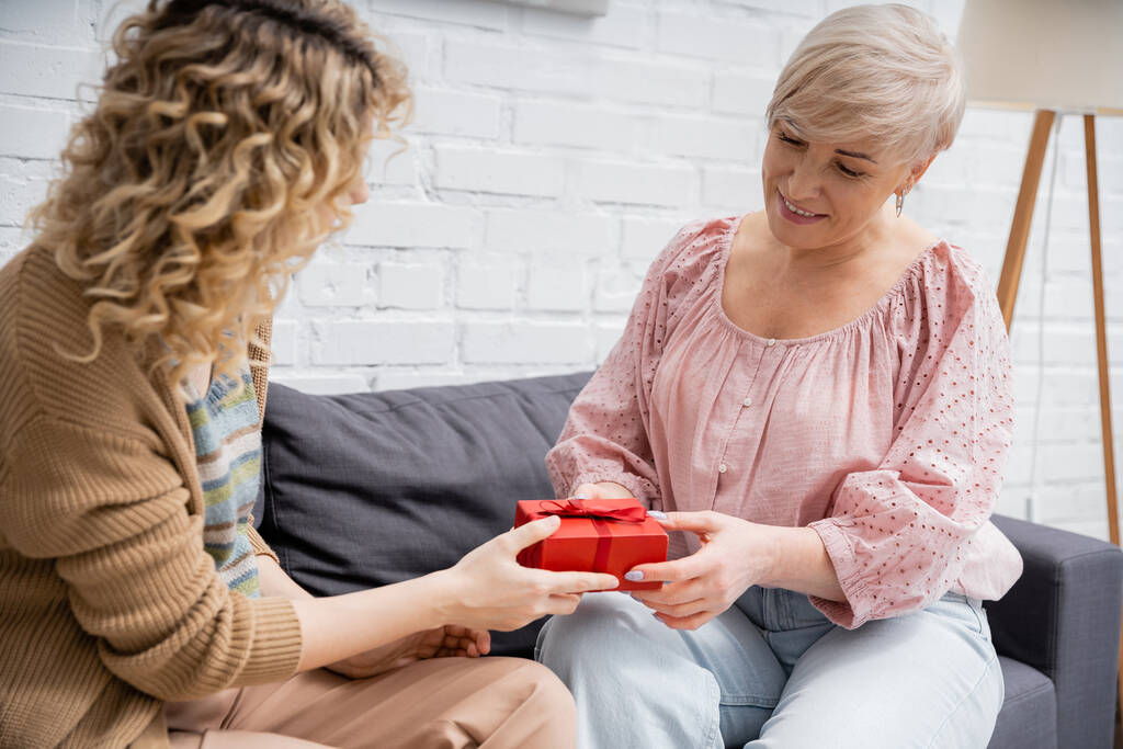 curly woman presenting gift to pleased mother while sitting on couch at home - Photo, Image