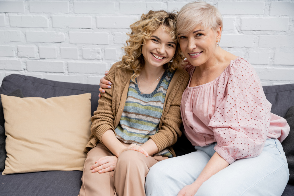 gelukkig volwassen vrouw met vrolijk blond dochter zitten op bank in woonkamer en kijken naar camera - Foto, afbeelding