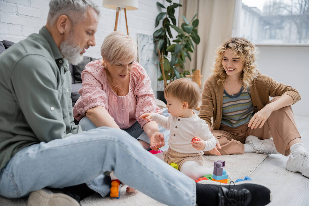 freudige Frau schaut Eltern mittleren Alters beim Bauklötzchenspiel mit Kind auf dem Fußboden im Wohnzimmer zu - Foto, Bild