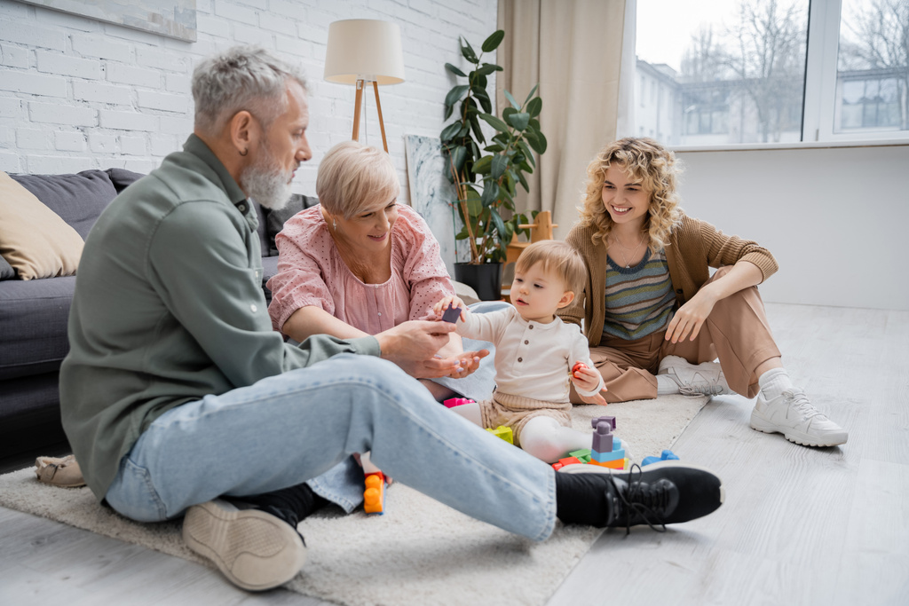 fröhliches reifes Paar spielt Bauklötzchenspiel mit Enkelin, während es im modernen Wohnzimmer auf dem Boden sitzt - Foto, Bild