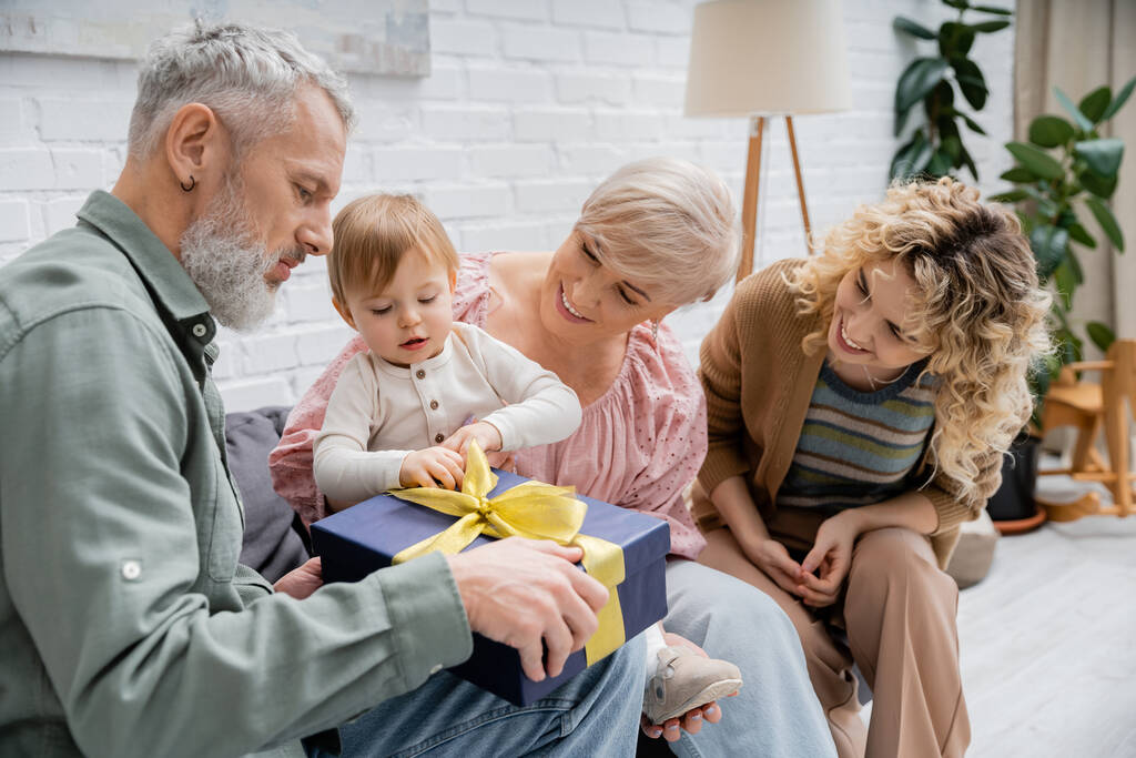 zralé vousatý muž držení dárek v blízkosti batole vnučka a usmívající se rodina v obývacím pokoji - Fotografie, Obrázek