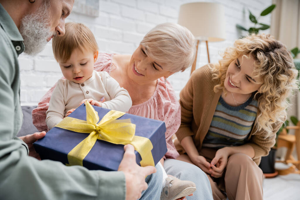 Bärtiger Mann hält Geschenkbox in der Nähe von Enkelin und Familie im Wohnzimmer - Foto, Bild
