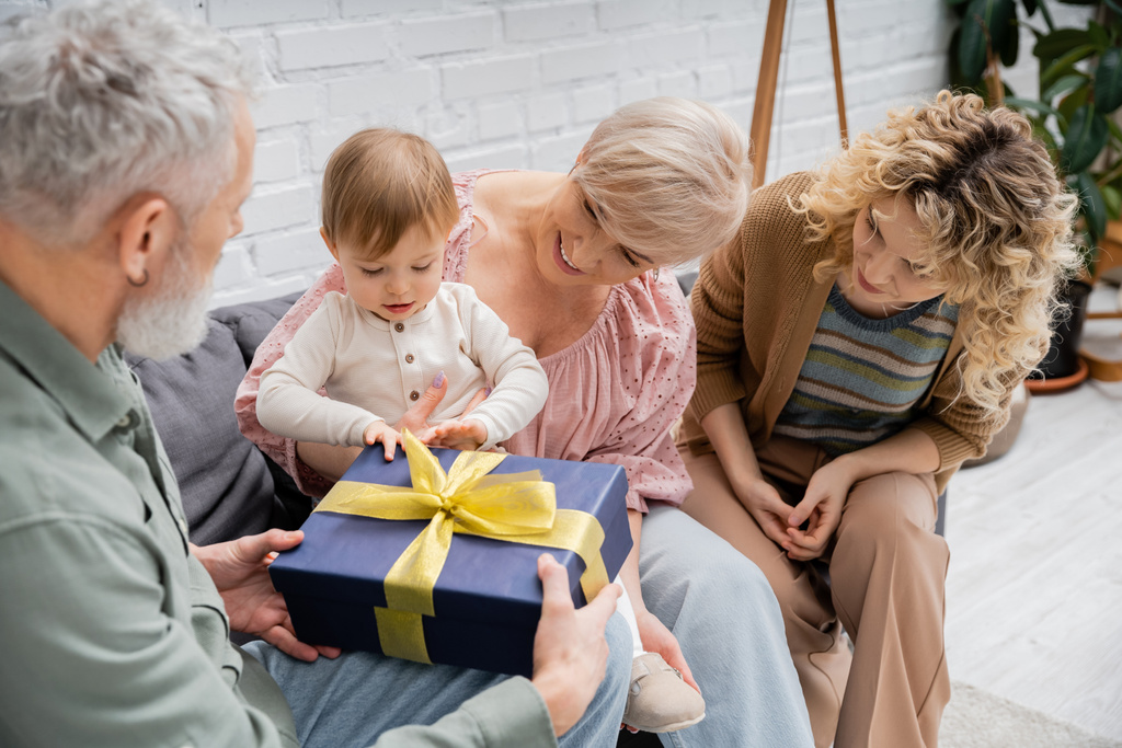 peuter kind opening geschenkdoos in de buurt van glimlachende moeder en grootouders op de bank in de woonkamer - Foto, afbeelding
