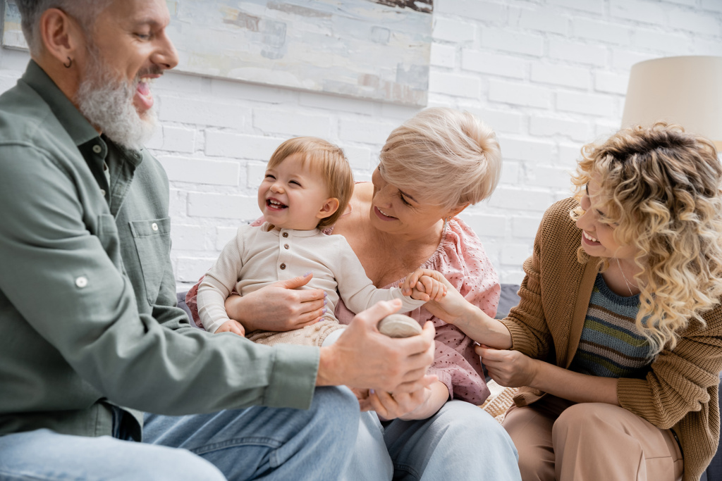 man met baard aanraken been van overgelukkig kleindochter terwijl plezier met familie op de bank in de woonkamer - Foto, afbeelding