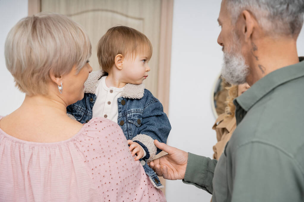 vousatý muž dotýkající se ruky malé vnučky v džínové bundě v blízkosti rodiny a vstupní dveře bytu  - Fotografie, Obrázek