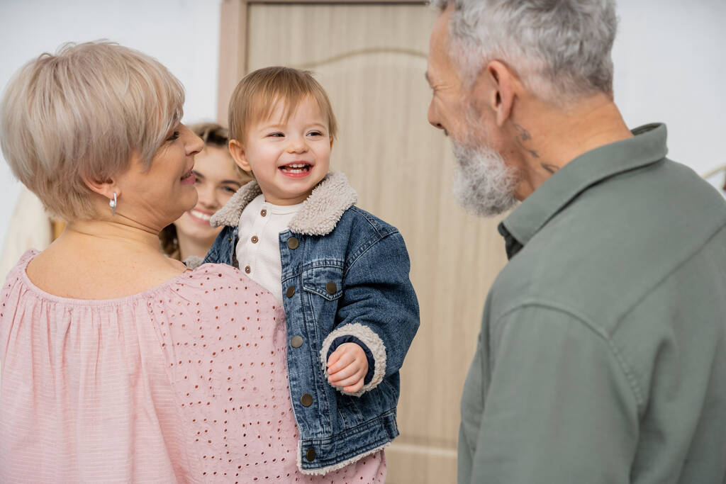 niño despreocupado en chaqueta de mezclilla sonriendo cerca de la madre y abuelos felices en el pasillo en casa - Foto, Imagen