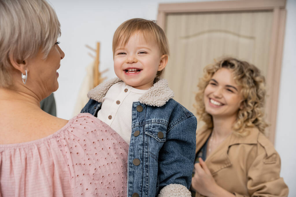 joyful child in denim jacket smiling in hands of granny near happy mother and entrance door on blurred background - Photo, Image