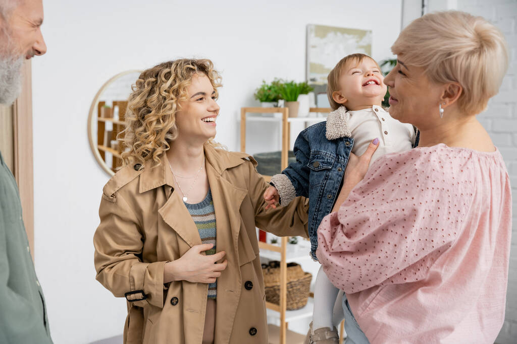 fröhliche Frau mittleren Alters hält Enkelin in Jeansjacke neben Familie im Wohnzimmer - Foto, Bild