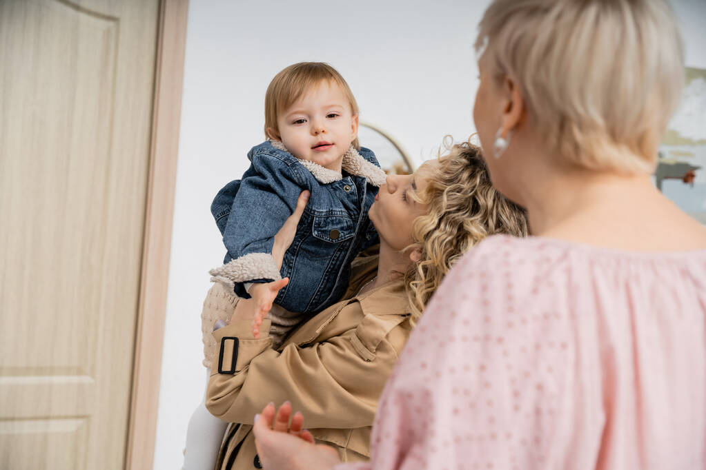 blurred middle aged woman looking at daughter holding little girl in denim jacket near entrance door of apartment - Photo, Image
