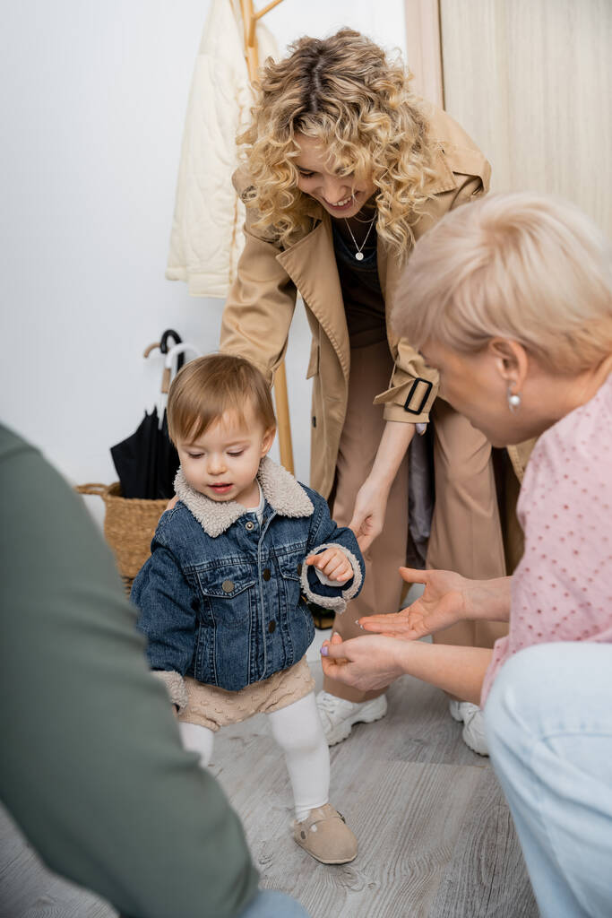 cheerful woman in trench coat and little child in denim jacket near blurred grandparents in hall at home - Photo, Image