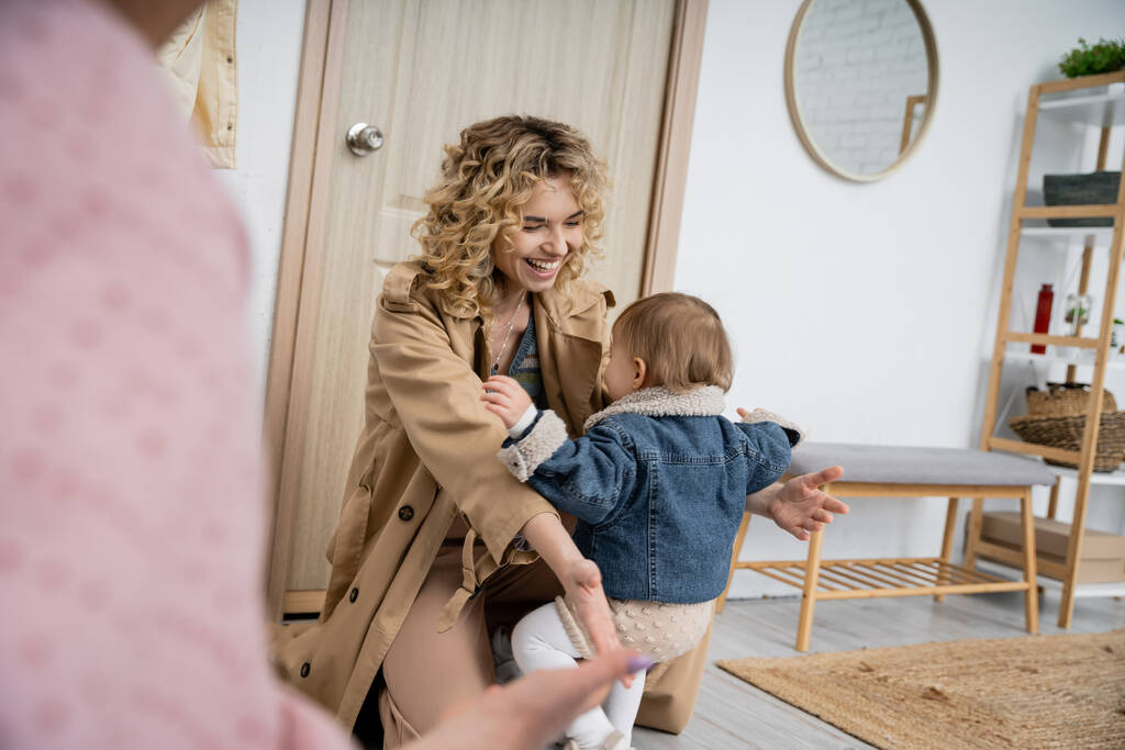 joyeuse femme en trench coat rencontre tout-petit fille près de la porte d'entrée à la maison - Photo, image