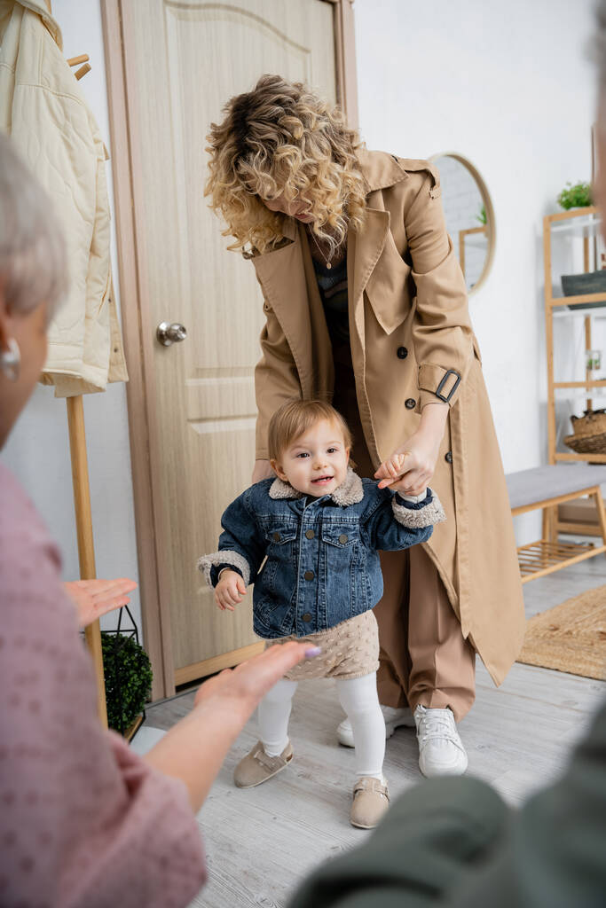 kobieta w płaszczu i dziewczynka w dżinsowej kurtce odwiedza dziadków w domu na niewyraźnym pierwszym planie - Zdjęcie, obraz