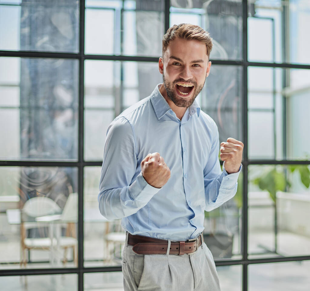 Πορτρέτο ενός νεαρού επιχειρηματία με μπλε πουκάμισο που κουνάει τις γροθιές του γιορτάζοντας την επιτυχία του στο γραφείο - Φωτογραφία, εικόνα