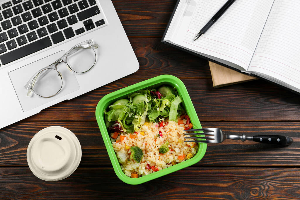 Container with tasty food, fork, laptop, paper cup of coffee and notebook on wooden table, flat lay. Business lunch - Photo, Image