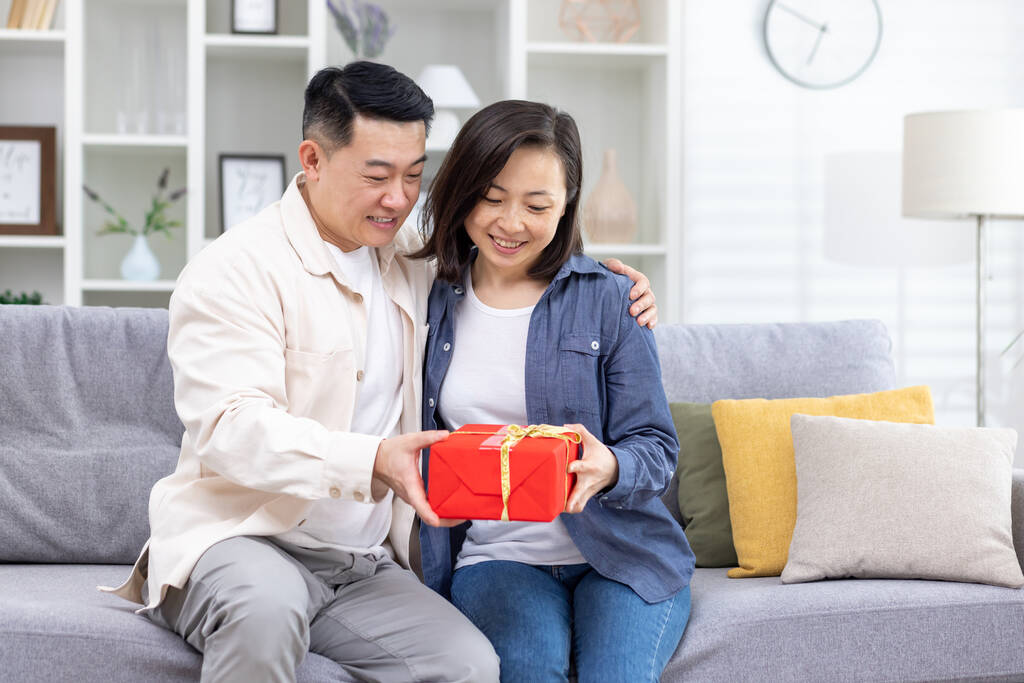 Joyeux jeune couple asiatique, homme et femme assis sur le canapé à la maison. Un homme donne à une femme une boîte rouge comme cadeau surprise. Ils se sourient.. - Photo, image
