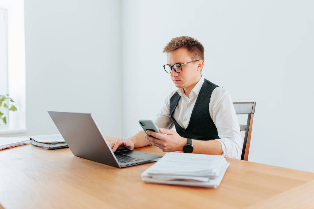 Ένας επιχειρηματίας, ένας τύπος σε ένα λευκό πουκάμισο κολάρο, χρησιμοποιώντας το smartphone του για να συνομιλήσετε με τον συνεργάτη του, ενώ εργάζονται για το laptop του στο γραφείο του στο σπίτι. - Φωτογραφία, εικόνα