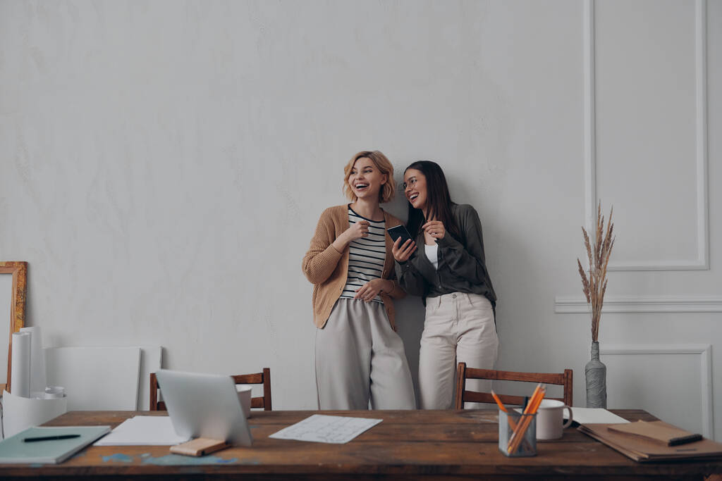 Zwei fröhliche junge Frauen unterhalten sich und lächeln, während sie in der Nähe ihres kreativen Arbeitsplatzes stehen - Foto, Bild
