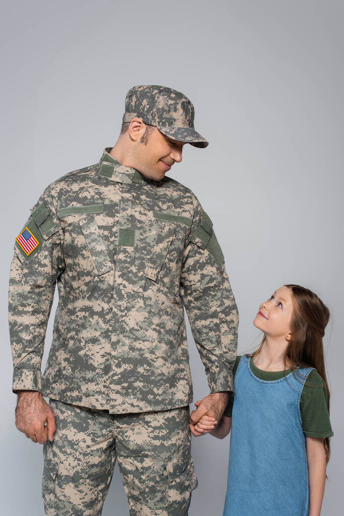ευτυχισμένος στρατιώτης με στρατιωτική στολή κρατώντας το χέρι με χαρούμενη κόρη κατά τη διάρκεια της ημέρας μνήμης απομονωμένη σε γκρι - Φωτογραφία, εικόνα