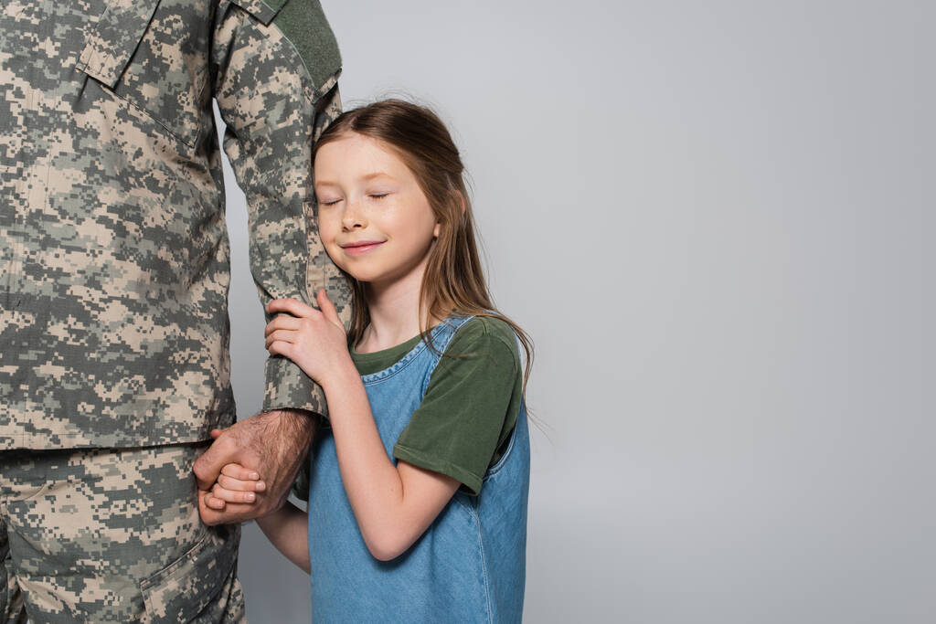 Asker üniformalı, babasının elini kucaklayan neşeli, çocuksu bir kız. Gri yolda izole edilmiş.  - Fotoğraf, Görsel