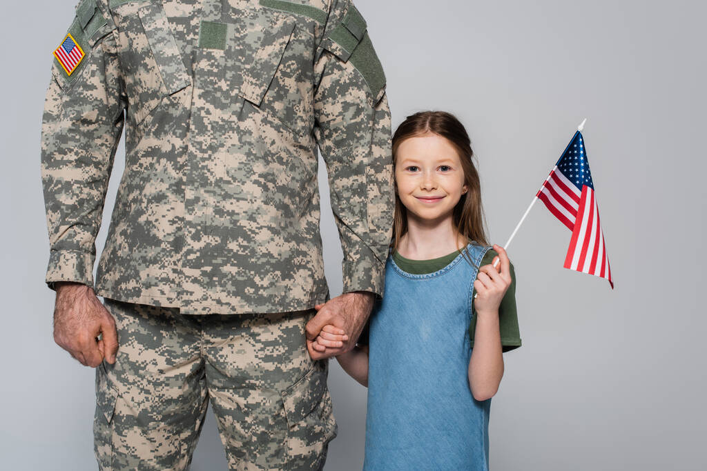 alegre niña preadolescente sosteniendo la bandera americana y la mano del padre en uniforme militar durante el día conmemorativo aislado en gris  - Foto, Imagen