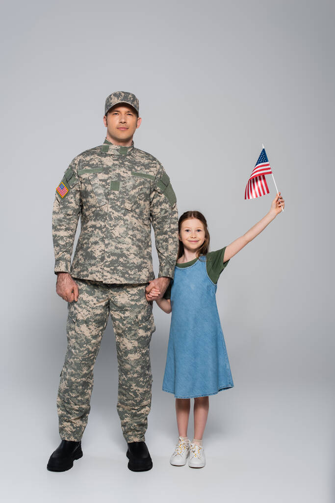 alegre niña preadolescente sosteniendo la bandera americana y la mano del padre en uniforme militar durante el día conmemorativo en gris  - Foto, Imagen